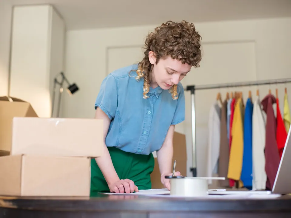 Online store owner packing customer orders into boxes beside a clothing rack